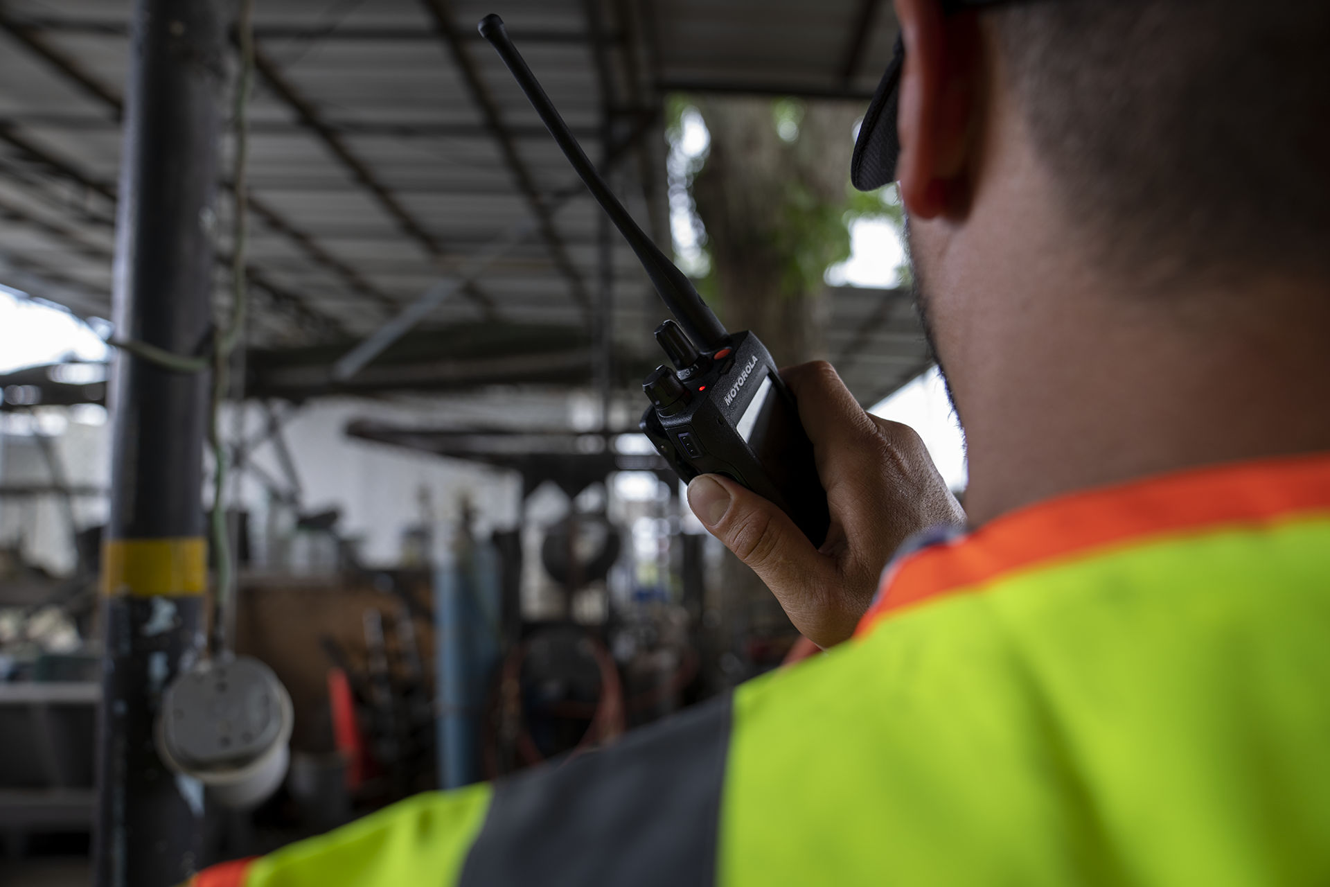 Worker in a neon safety vest uses a handheld two-way radio inside an industrial workshop.