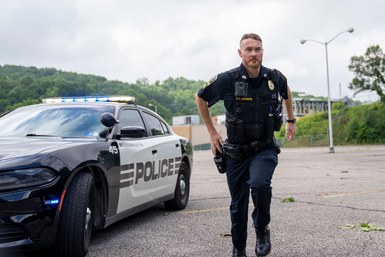 Police officer running past a black-and-white patrol car with blue lights in a parking lot.