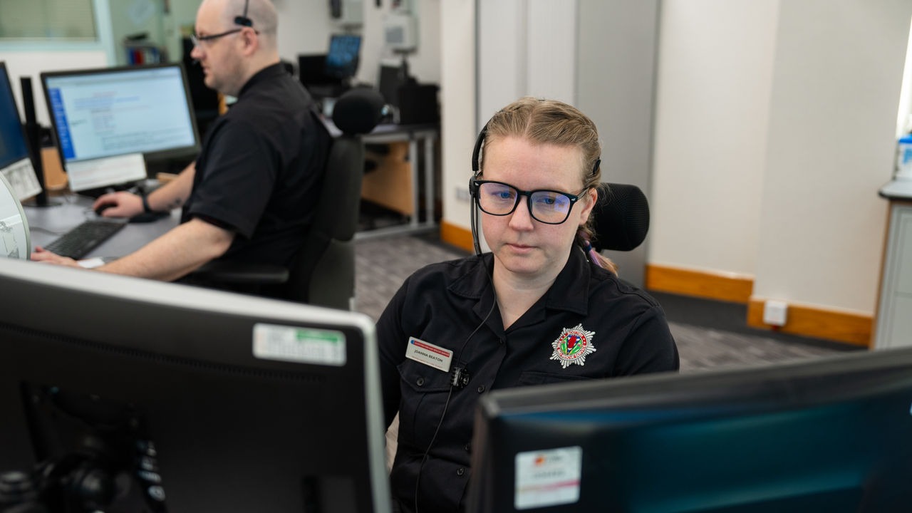 Two call-center operators in black uniforms wearing headsets at computer desks, focused on screens in a dispatch center (front-facing woman).