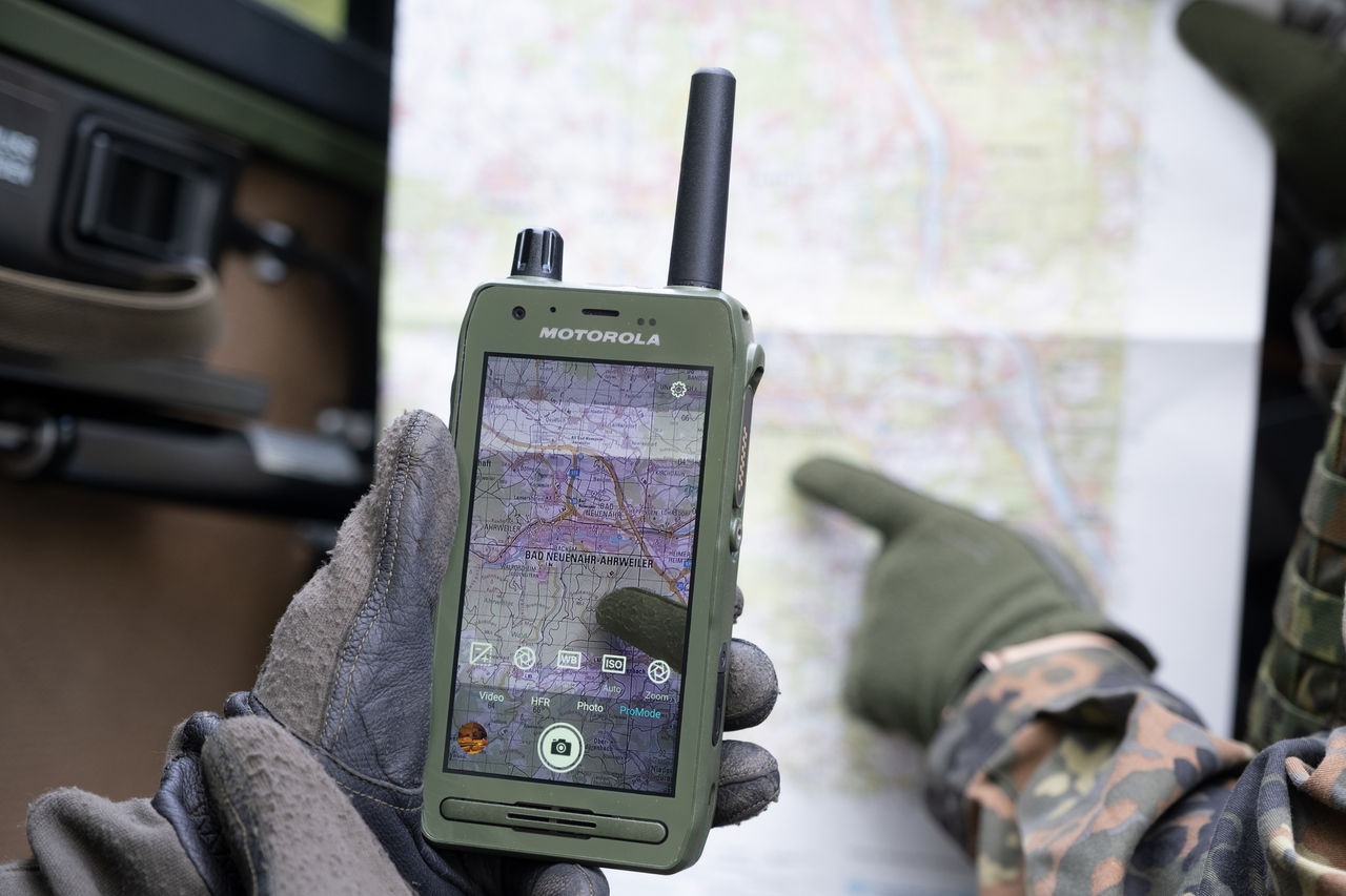 Rugged Motorola handheld radio with antenna, held by gloved hands over a map showing GPS navigation.