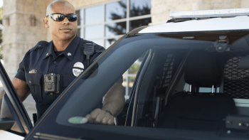 Police officer running past a black-and-white patrol car with blue lights in a parking lot.